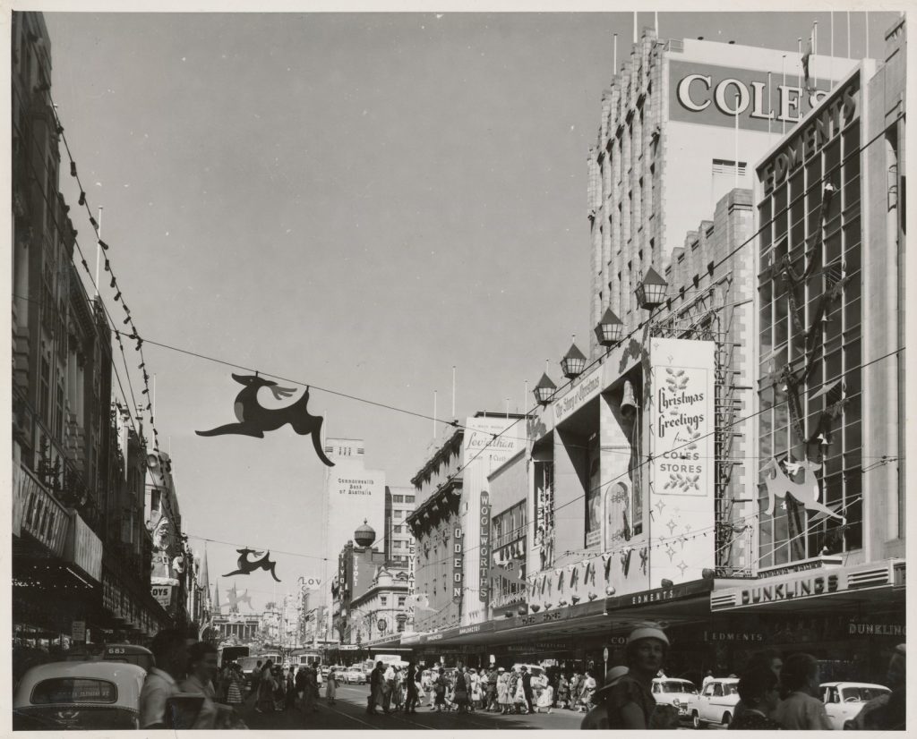 View of Bourke Street shipping strip with vehicles and people. Christmas decorations can be seen including reindeers suspenders from wires above the road. 