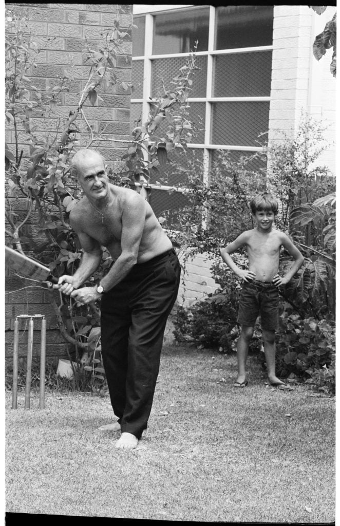 Shirtless man playing backyard cricket, batting in front of cricket stumps. Boy standing nearby observing.
