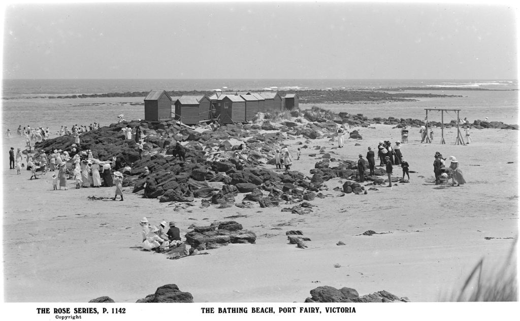 Rocky beach with a collection of small wooden buildings sitting on the rocks. People at the beach are fully dressed and wearing hats. 