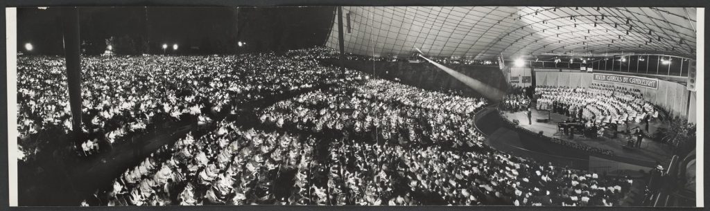 Elevated view of seated audience holding candles, and candle light from the rest of the audience seated on the lawns in background, lit stage with seated choir and musicians in right background