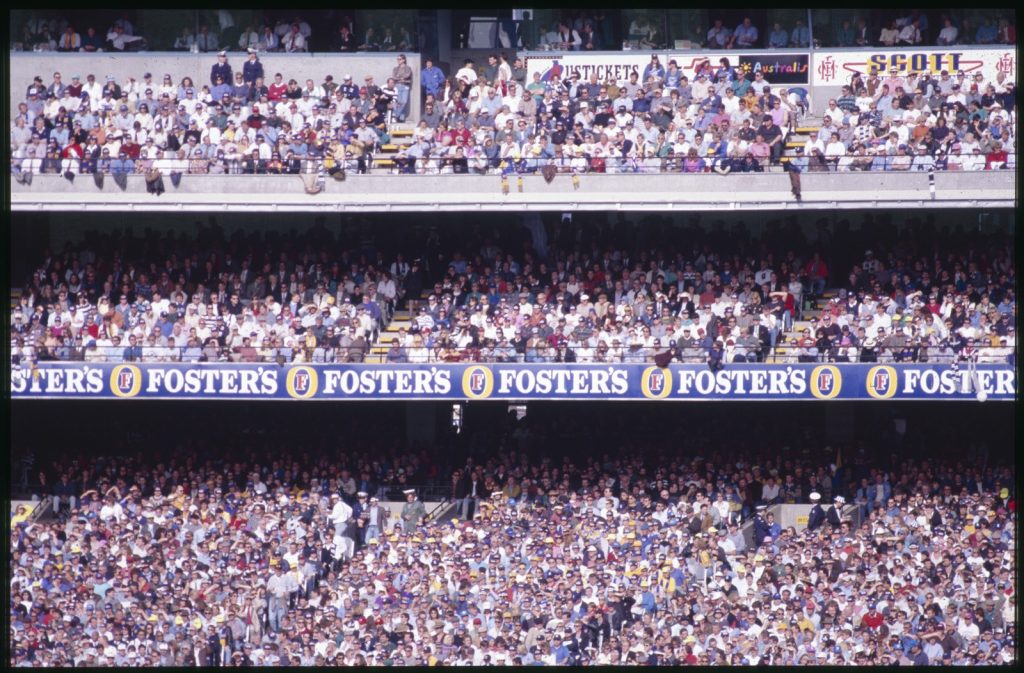 Crowds of people at the MCG in the grandstand; Foster's sponsorship.