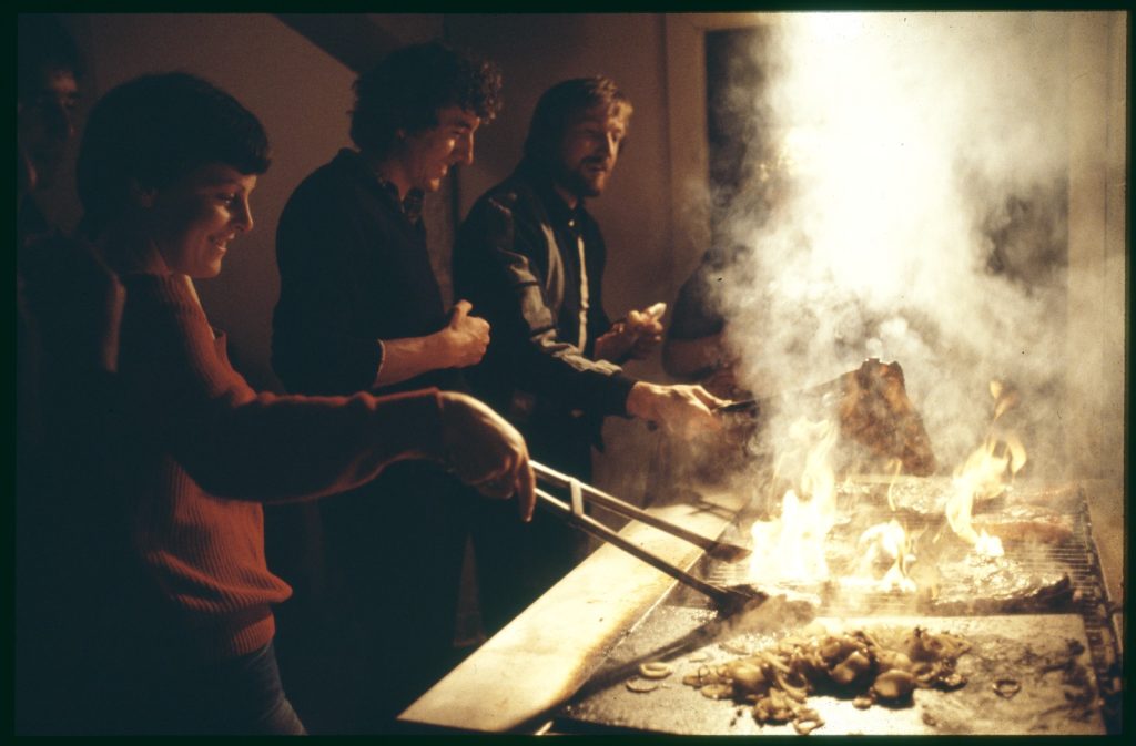 Three people in front of a smoking BBQ in dim light or twilight, cooking meat.  