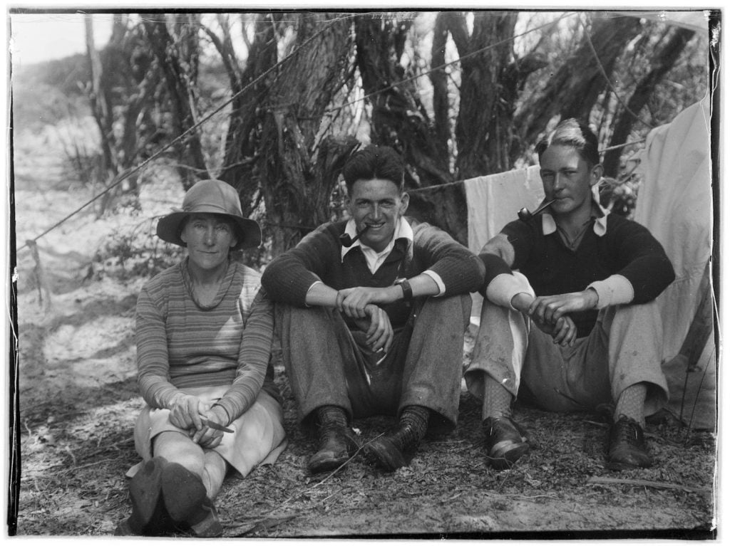 Woman and 2 men, sitting at the campsite in the shade with a line of towels behind them