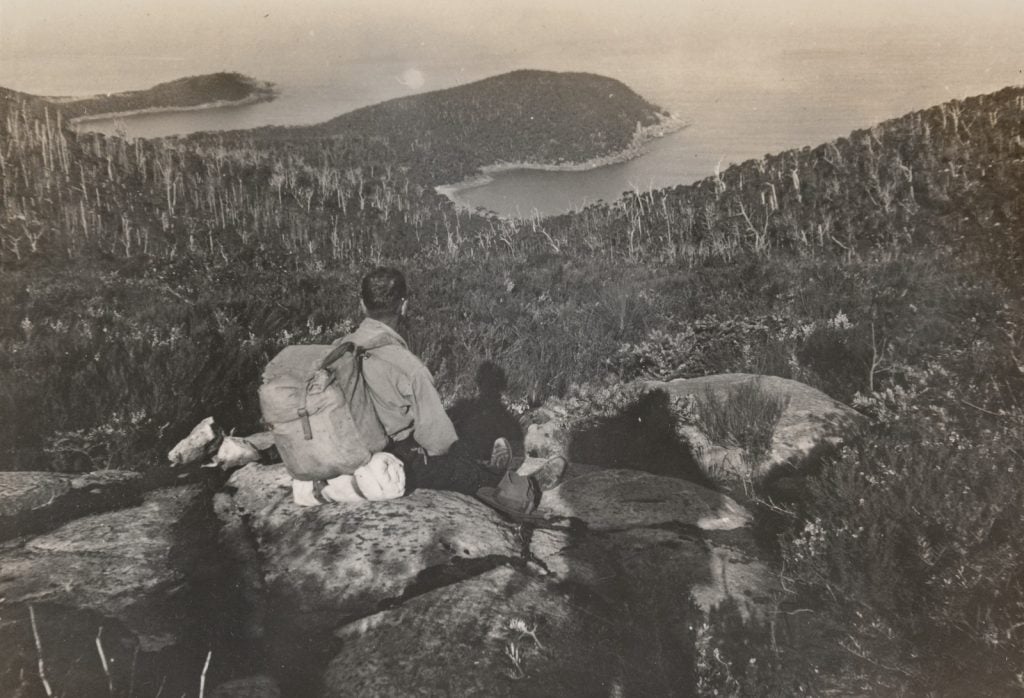 Man with a backpack sitting on top of a rocky outcrop overlooking recently burnt forest, down to the coast