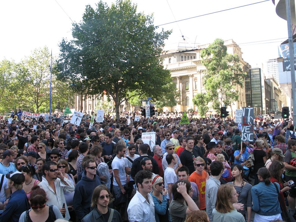 Colour photograph, slightly elevated view of large crowd of people, many holding placards, at a rally in front of State Library Victoria.