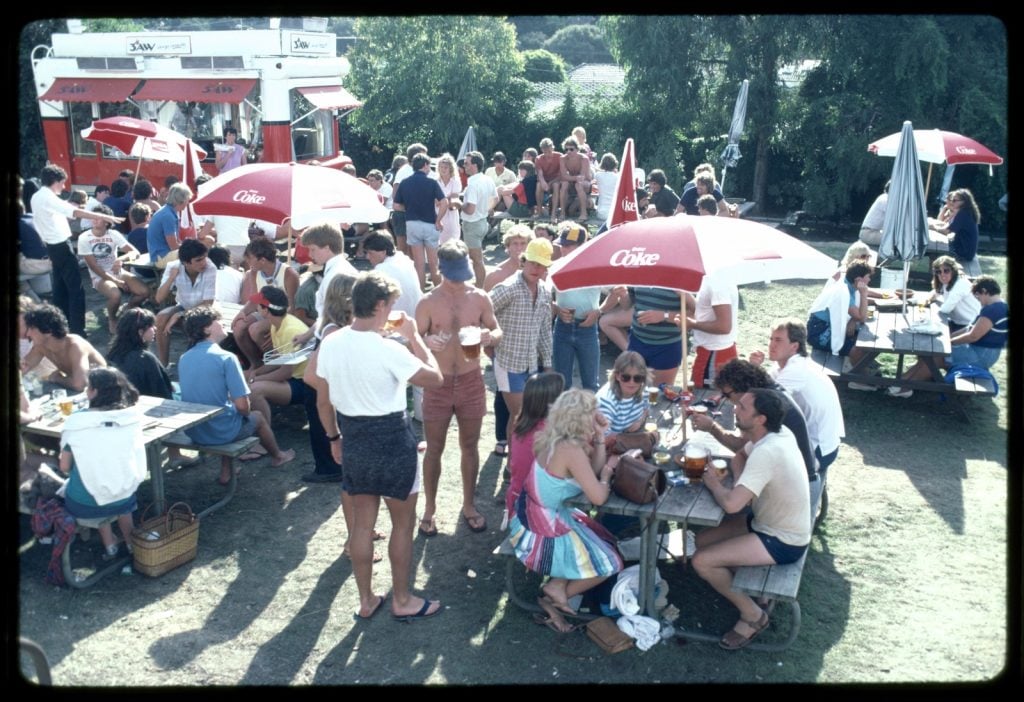 Whole-length group portrait of people seated at tables with red and white "Enjoy Coke" Umbrellas and other people standing around; also beer jugs and beer glasses and in the background the 3AW Radio station van.