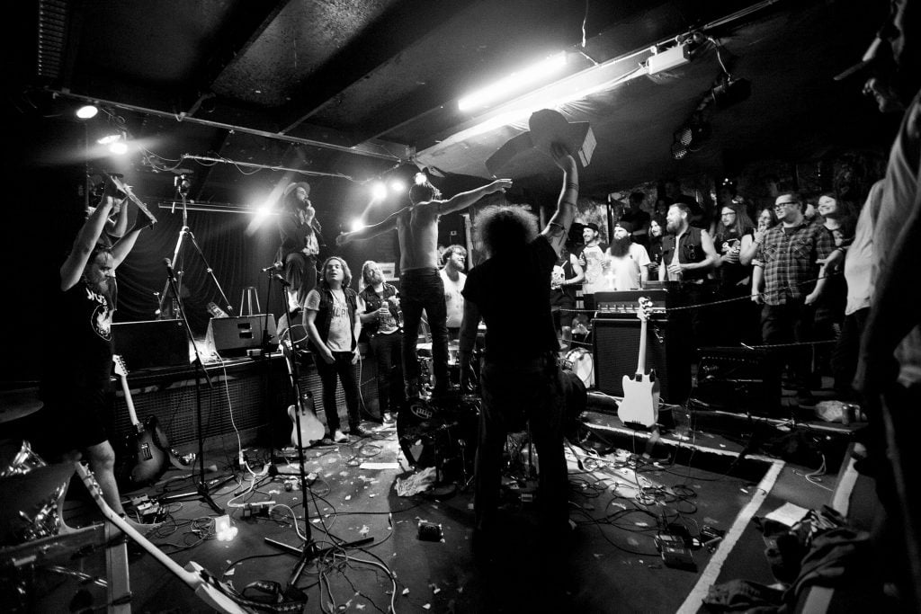 Black and white photograph of inside of music venue with several people holding guitars over their heads in a sunken dancefloor, many people watching behind.