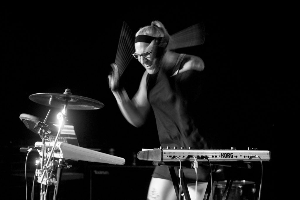 Black and white action photograph of standing person holding drumsticks playing electronic trigger pad and cymbals with Korg synthesizer in front.