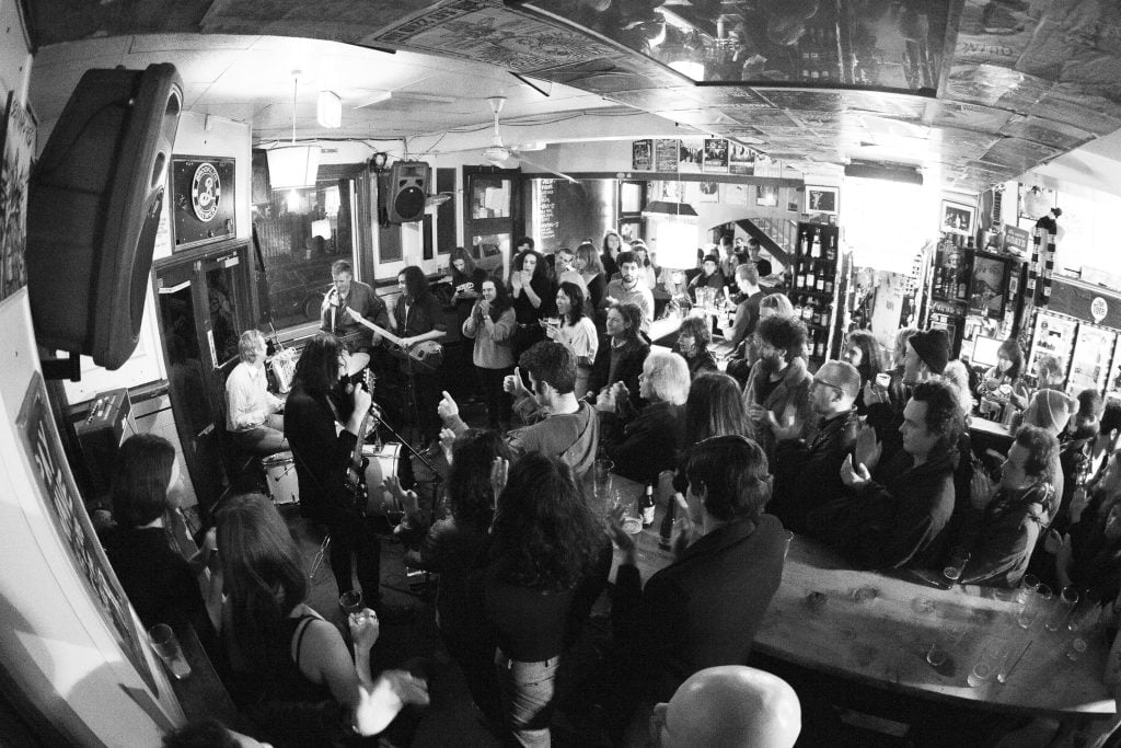 Black and white photograph, elevated with fish-eye lense. A crowd watches a four-piece band playing in corner of a bar.