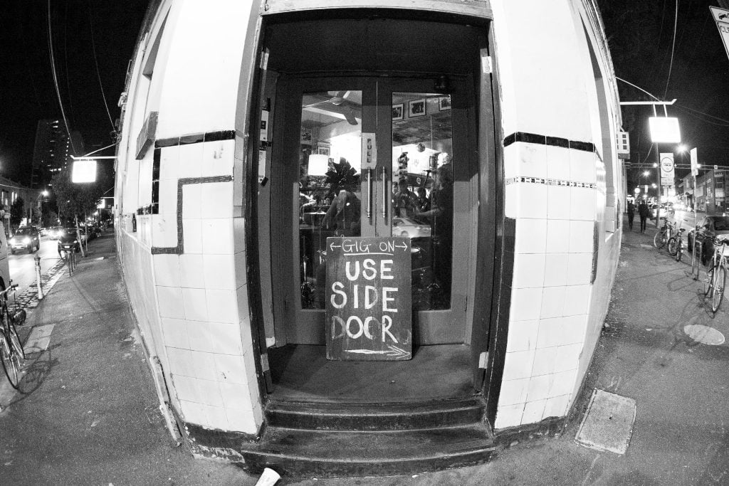 Fish-eye photograph of the corner entry to The Tote Hotel, Collingwood. Sign on front step reads 'Gig on use side door'.
