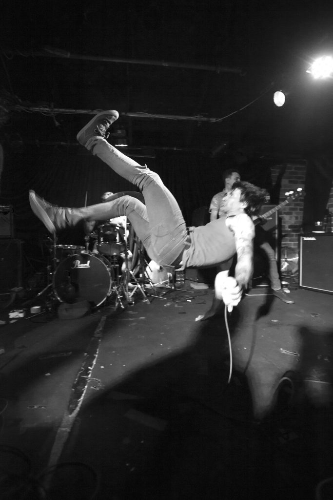 Black and white action photograph of person with microphone in hand, in mid-air, about to land on their back, presumably after falling or jumping backwards. Drummer and bass player in background.