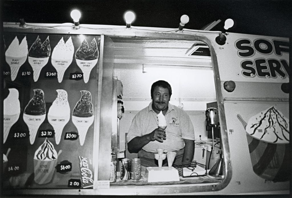 Black and white photo of vendor at the window of his ice cream van. He is holding a soft serve ice cream. 