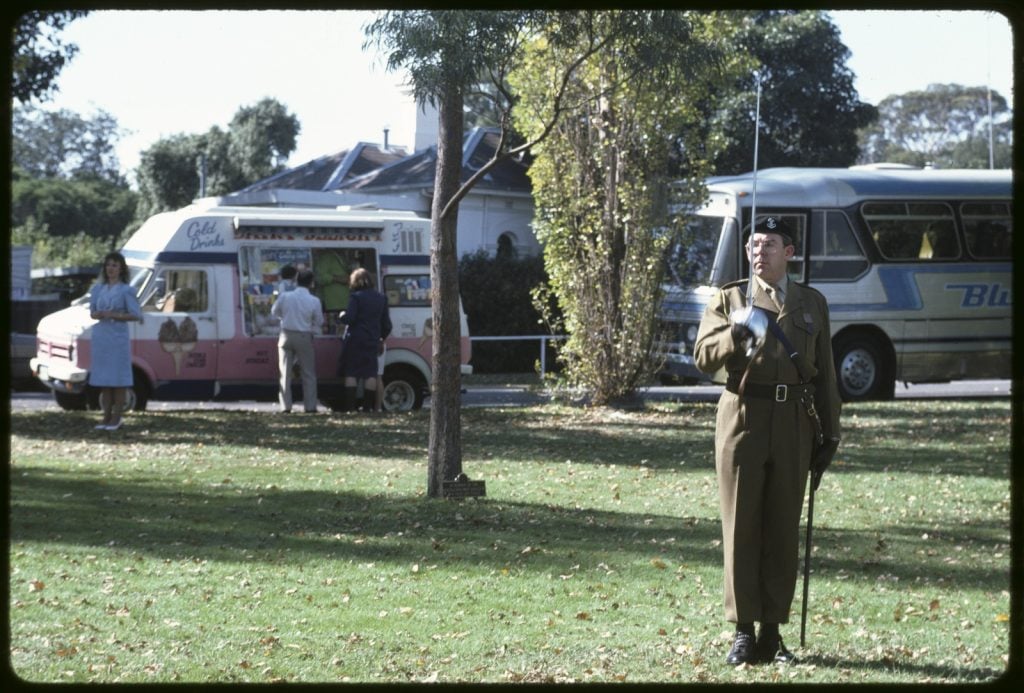 Photo of soldier standing to attention in the park, icecream vans sells ice creams in background. 