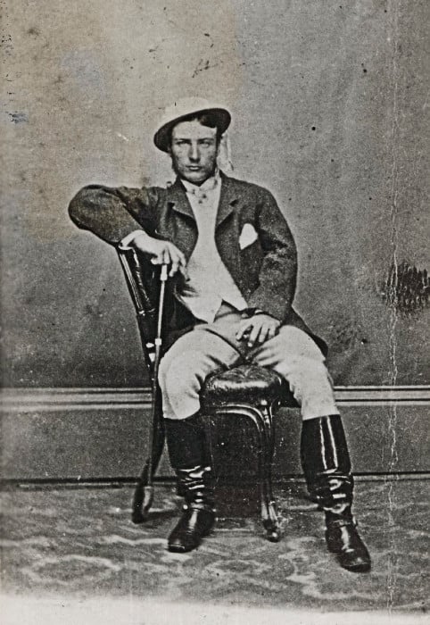 Whole-length black and white photo of Marcus Clarke sitting sideways on a chair, with his right arm resting on the chair back. He is holding a cane in his right hand and wearing a hat.