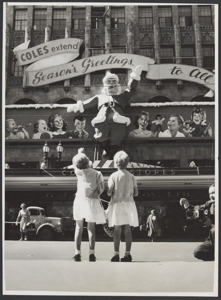 Two girls in dresses with their backs to the camera  looking at Christmas decorations on the Coles Bourke Street Store No. 12, which includes a large Santa figure. 