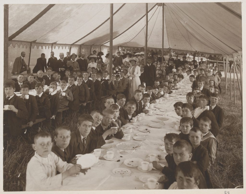 Large group of boys sitting at two long tables inside a large marquee. White table cloths, cup and saucers and plates are on the tables.  