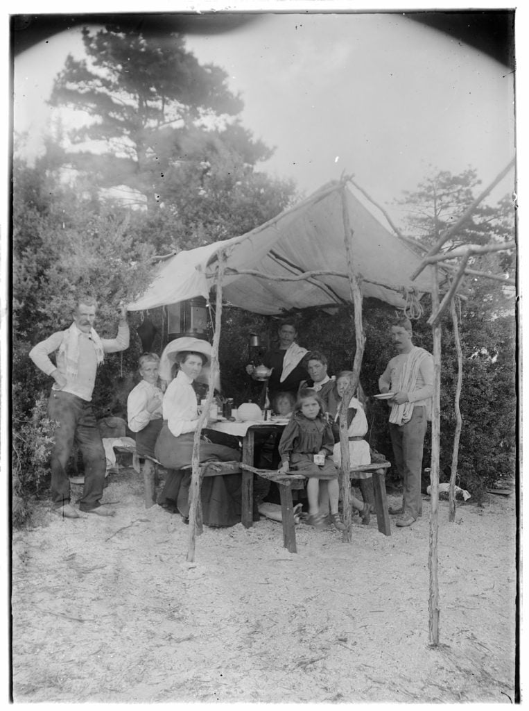 Family group at picnic on beach. Women and children sit at picnic table under makeshift roof.  Three men stand beside table. 