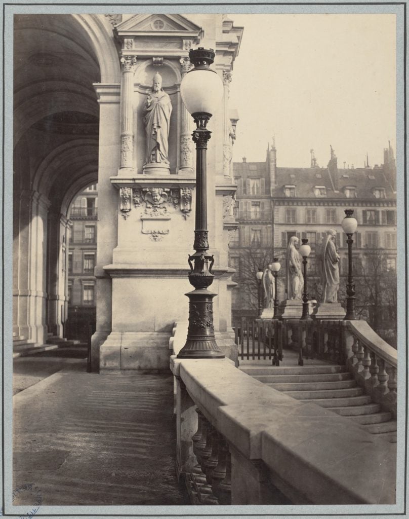 Single globe with iron lamp post mounted on marble top of a balustrade. Statue in niche in background and others along balustrade in right background.