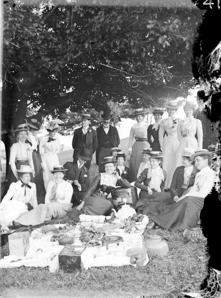 Group of men and women at a picnic. Some sit and some stand. All wear hats. There is a cloth  covered in food and a large teapot. They look happy.