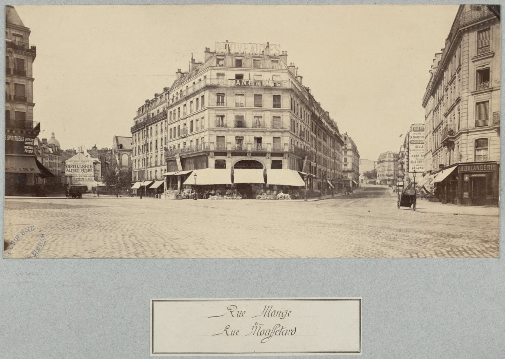 Looking from intersection of two streets with view along both streets showing corner building in centre signed "Augrand Monge" with goods displayed under awning over footpath, corner buildings left and right, restaurant and bakery.