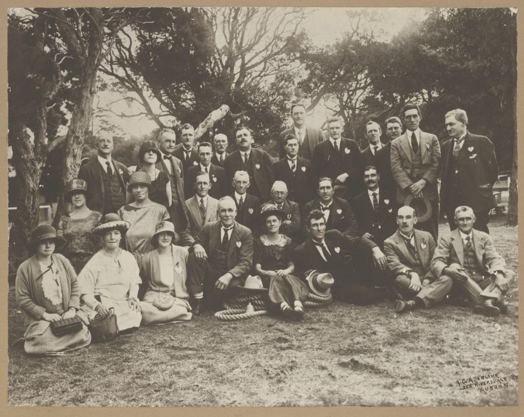 Large group of men and women, men dressed in suits, sitting and standing outdoors. Trees in background. 