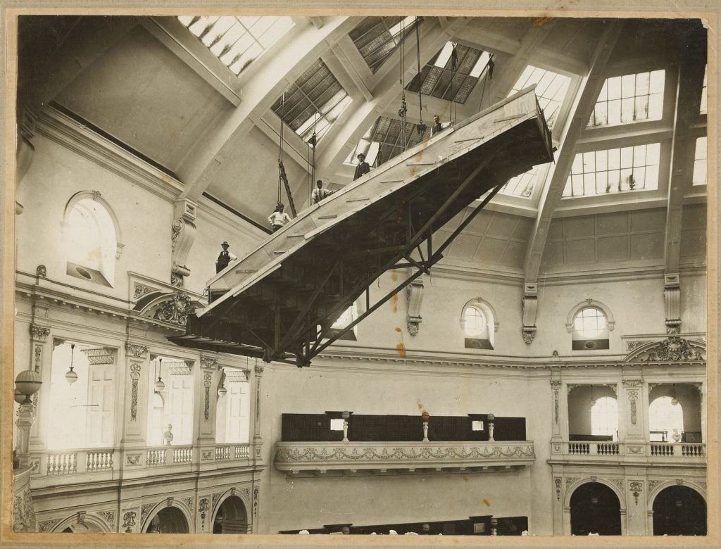 Interior of the Domed Reading Room, showing men standing on a portable scaffold, suspended from the ceiling, to enable repairs to the glass skylights