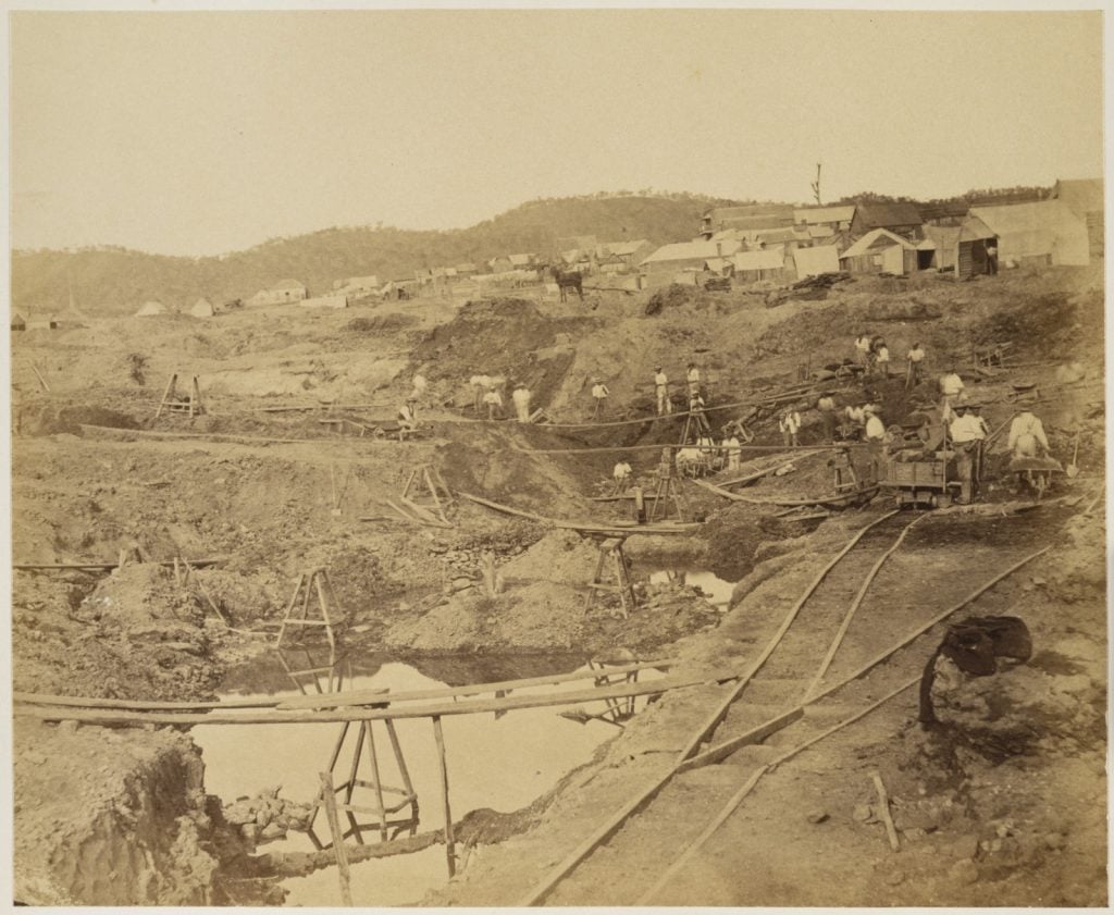men working at a goldmine along a creek with a train line for carting ore and buildings on the hill behind.
