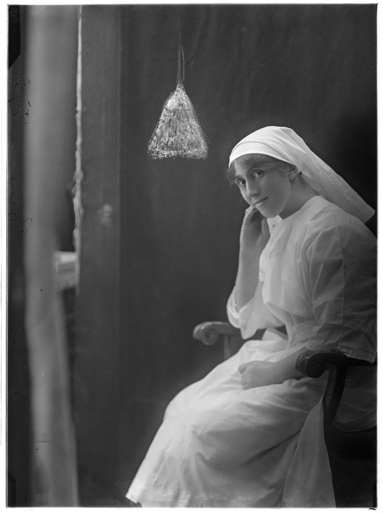 Photographic portrait of a nurse wearing a white uniform and and cap and scarf head covering, looking wisfully sideways at the camera 