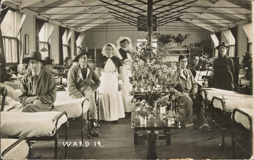 Nurses and patients in a ward, with long narrrow tables of flowers down the centre of the ward, 