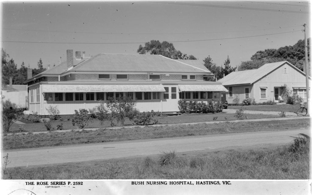 Single storey hospital buildings, on a dirt road with gardens around.