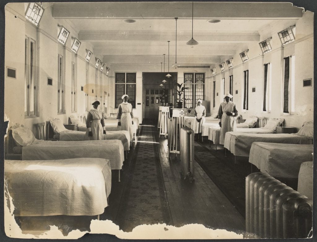 a ward with nurses standing beside beds, a row of large radiator heaters and table with plants down the centre of the room