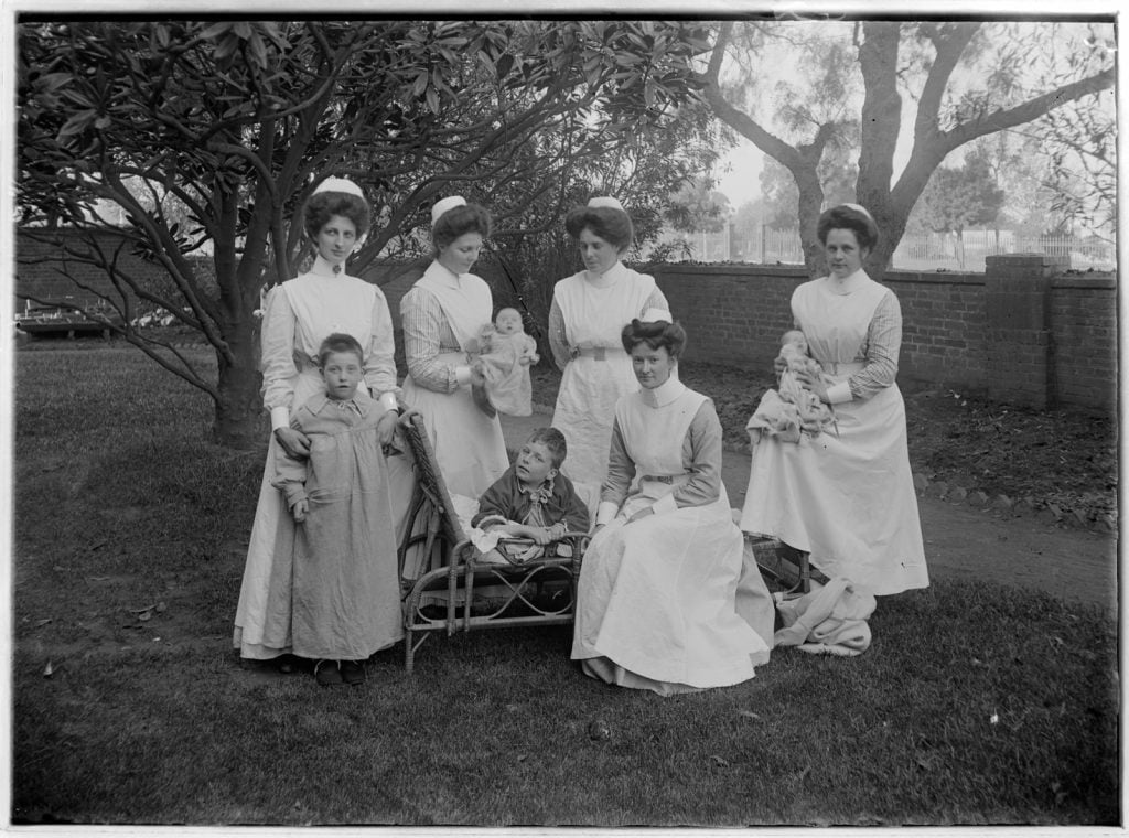 photograph of nurses, babies and children sitting outside, some on a cane lounge. Nurses wearing full length uniforms with caps, aprons and collars.