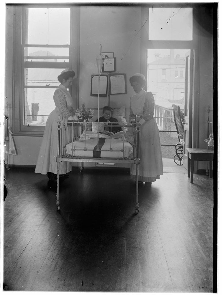2 nurses beside a child in a bed, the the windows and door to the outside are open