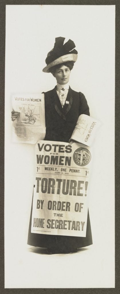 Black and white photo of Vida Goldstein holding a placard that reads: "Votes for Women - Torture! By order of the Home Secretary". The placard is dated 28 June 1912. She also holds copies of the suffragette newspaper, 'Votes for Women',
