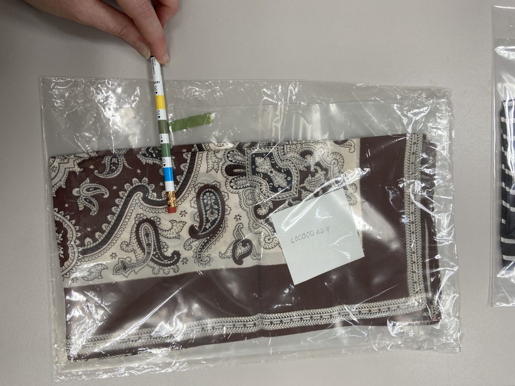 A brown and white vintage scarf inside a clear plastic enclosure with a coloured test paper and indicator pencil 