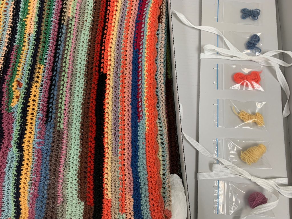A blue-grey board archival box containing a handmade, multi-coloured blanket on the left and leftover threads in little zip-lock bags on the right.
