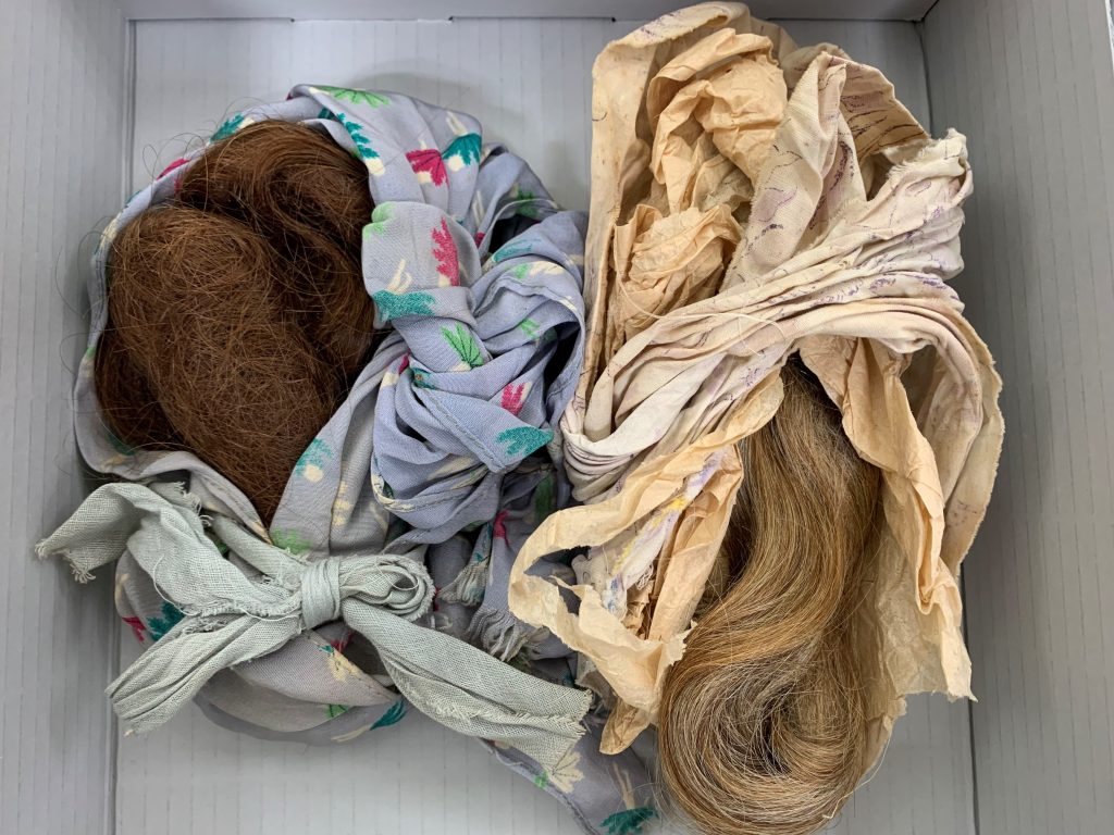Two bundles of women'd hair wrapped in vintage fabrics inside a blue-grey board archive box