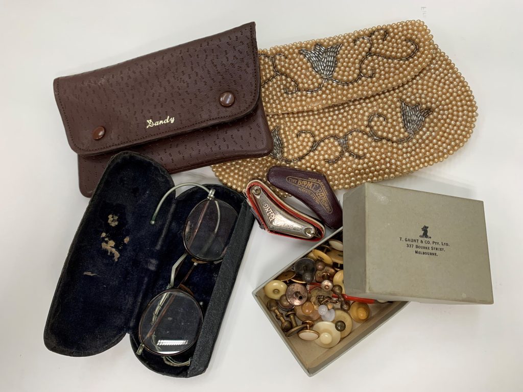 An assortment of vintage items on a white tabletop, including a pair of old spectacles in an open case, a brown leather wallet, a cream-coloured beaded purse and an open cardboard box of cufflinks