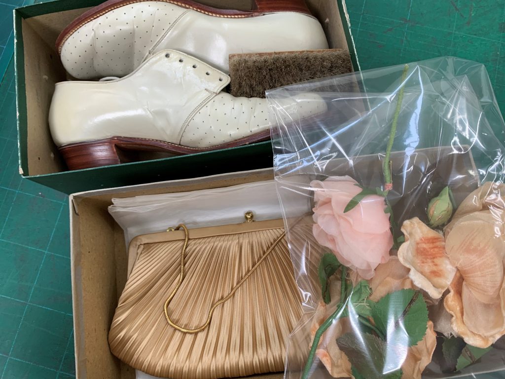 Two old shoeboxes. One contains a pair of white, vintage leather shoes. The other contains a gold pleated vintage handbag. There is a clear plastic bag containing apricot and light pink artificial roses to the side of the image.