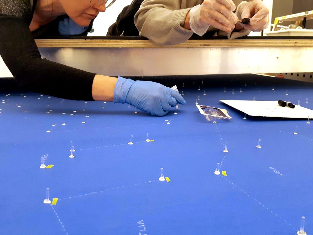 Two people with gloved hands lean over a moveable gantry structure, as they adhere small rods of clear acylic onto a sheet of blue fabric.
