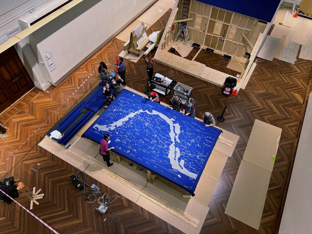 Aerial view looking down onto a large, knitted map of the stars in a gallery. The map is largely knitted in blue wool.