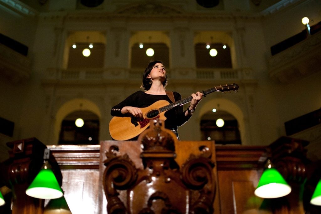 Photograph looking slightly up towards singer-songwriter Missy Higgins, a woman with short dark hair, holding an acoustic guitar. She is standing on the Dais in the Dome, and appears to be singing.