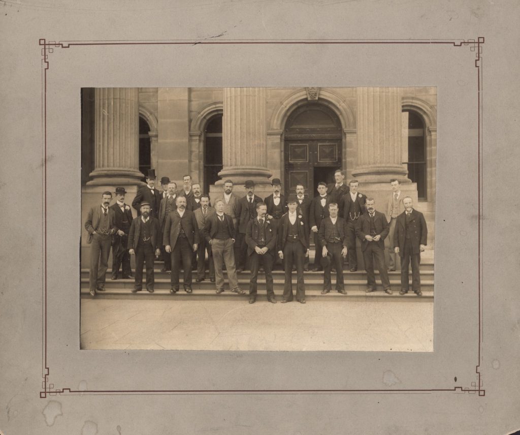 Two rows of men standing on front steps of library, back row: Armstrong Boys, Harvey Weatherall, Quirk, Bryant, Shellew, Vogler, O'Malley, Irving, [unnamed], Atkinson; front row: Kerr, Walcott, Barbey, Simpson, Mattham, Edwards, J. E. Shield, [unnamed], Dymond.
