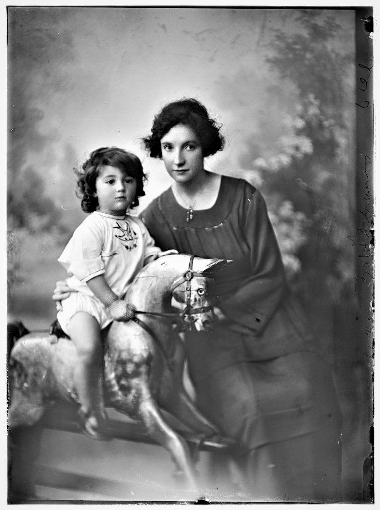 young woman with a bobbed haircut, next to a young child on a rocking horse. 