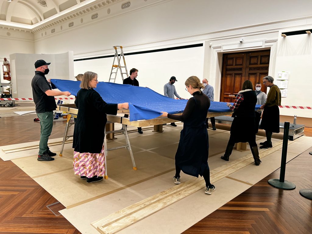 A group of people collectively lift a large rectangle of blue fabric above a raised wooden frame in a gallery space.