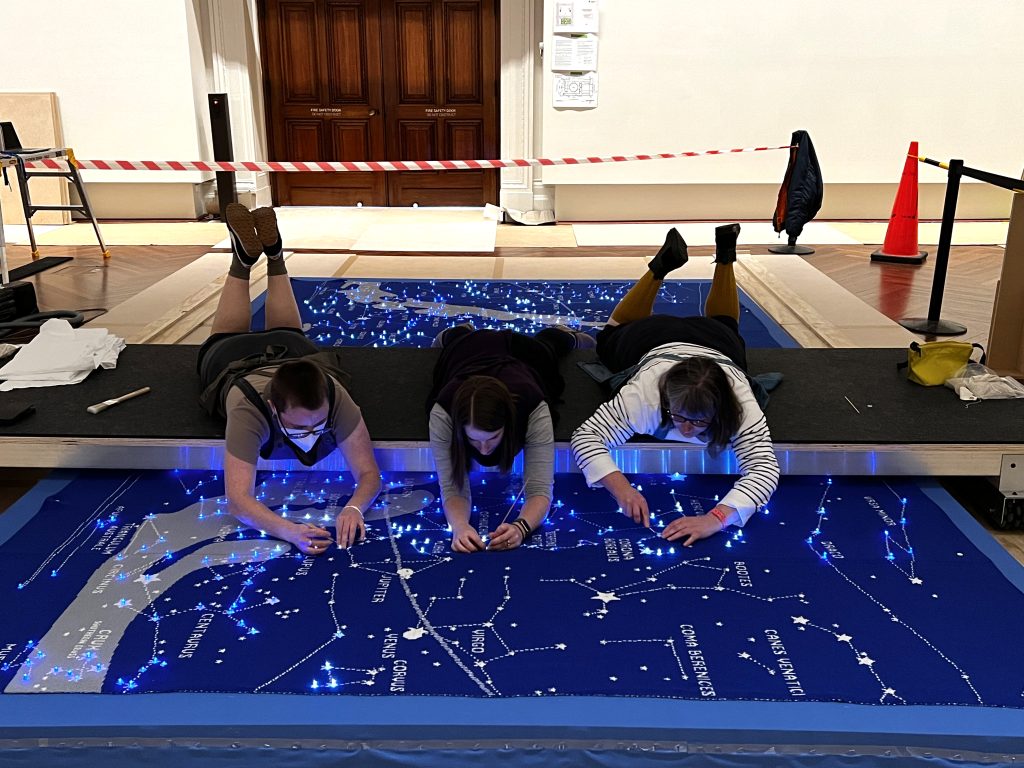 Laying on their fronts on a moveable gantry, three people look down toward a large blue knitted map of the stars. Some of the constellations are lit up.