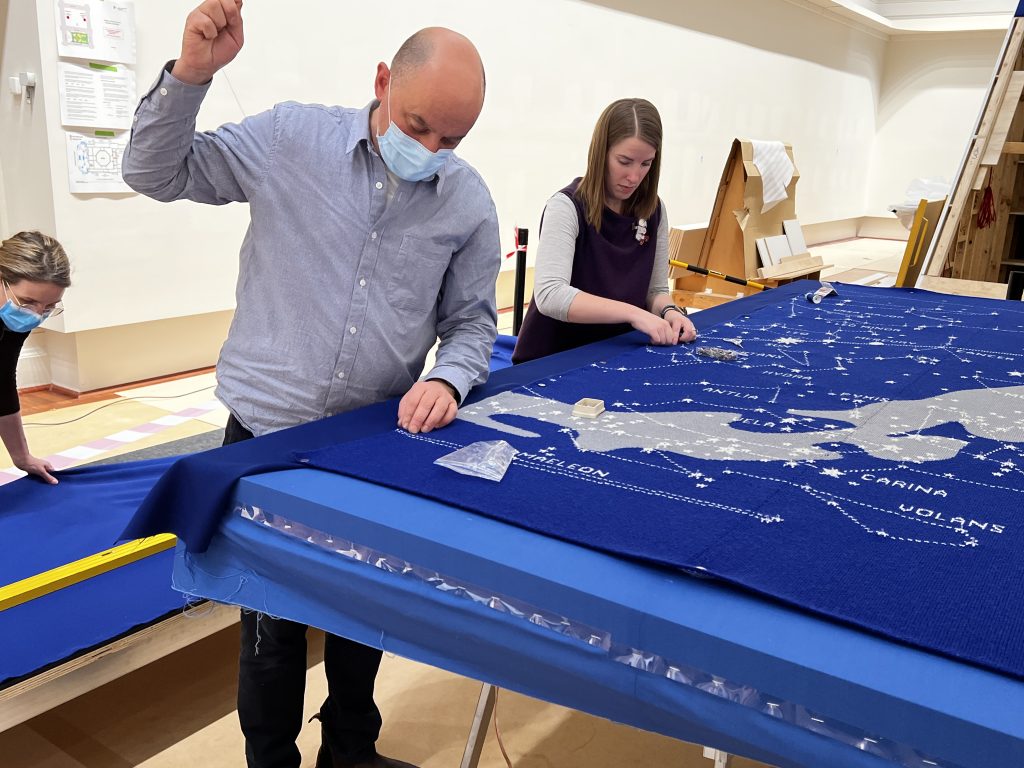 Two people stand at the edge of a large blue knitted map of the stars, in the process of sewing the map in place on a frame. A third person wears a mask at the far left on the frame.