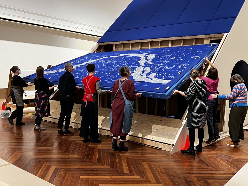 A group of people lift a large blue knitted map of the stars into a display structure in a gallery space.
