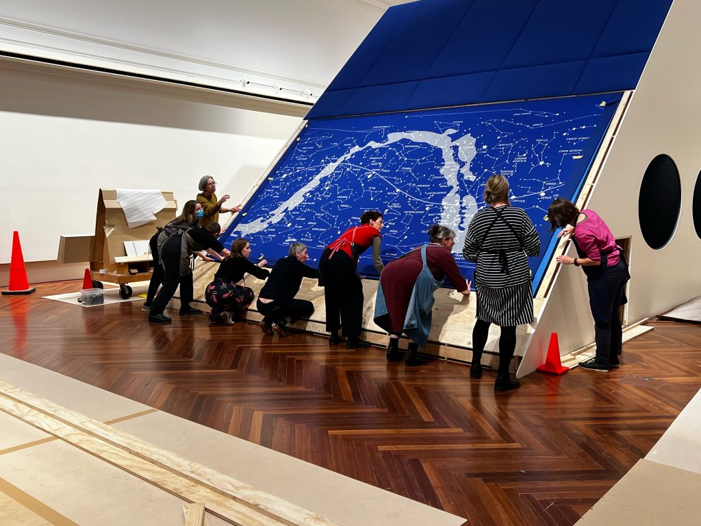 A group of people check the position of a large blue knitted map of the stars that has been inserted into a display structure in a gallery space.