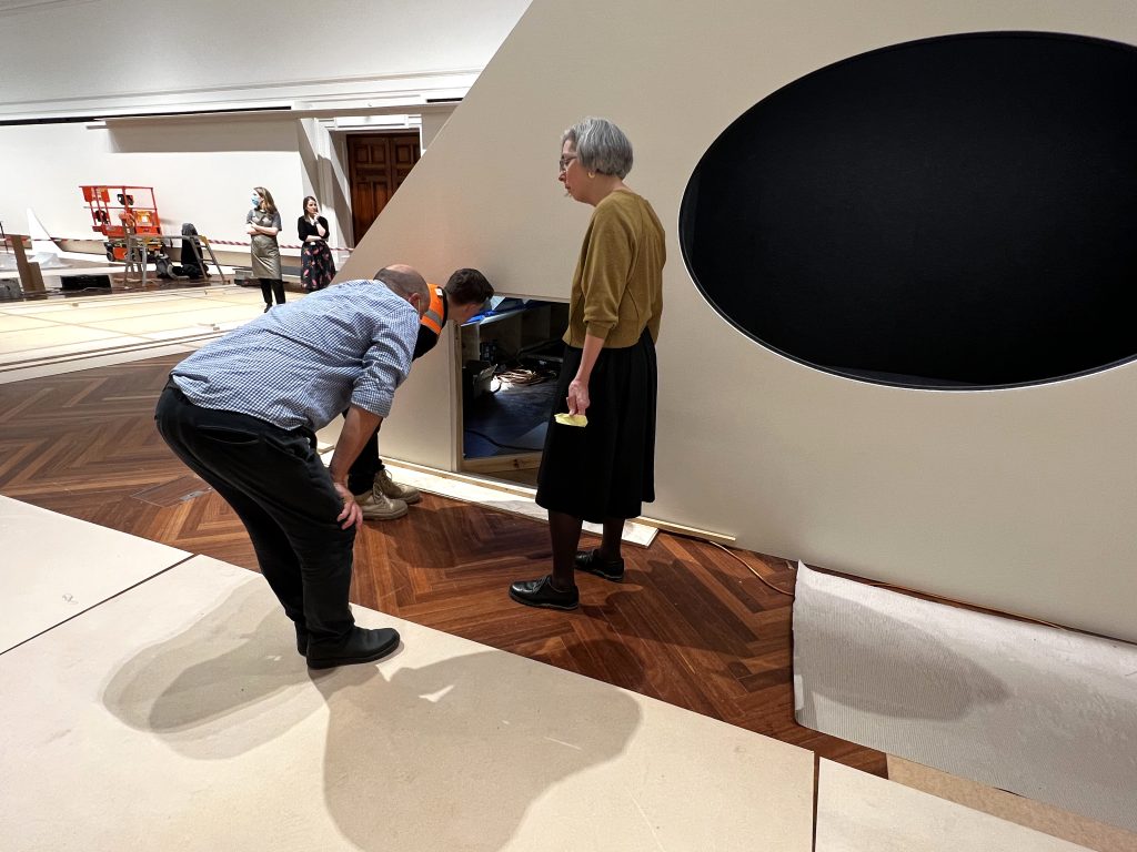 Two people are bent down, looking into a small doorway built into a large display structure in a gallery. A third person is standing next to them, observing.
